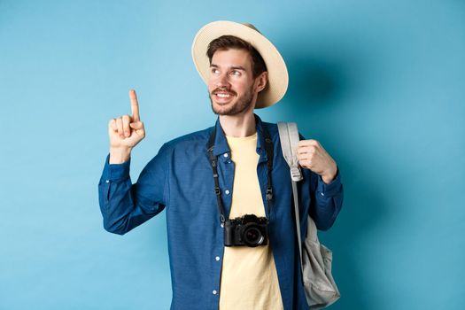 Handsome positive guy on vacation, wearing summer hat, looking and pointing up at logo, smiling pleased, standing with camera and backpack on blue background.