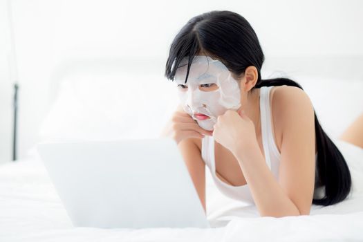 Beautiful young asian woman with sheet facial mask lying working on laptop computer on bed at bedroom, beauty girl applying face mask for skincare of wrinkle at home, skin care, health and wellness.