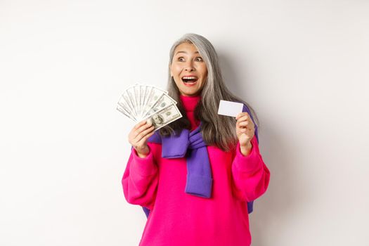 Shopping concept. Happy asian old woman with grey hair, looking fascinated at upper left corner, showing plastic credit card and money dollars, white background.