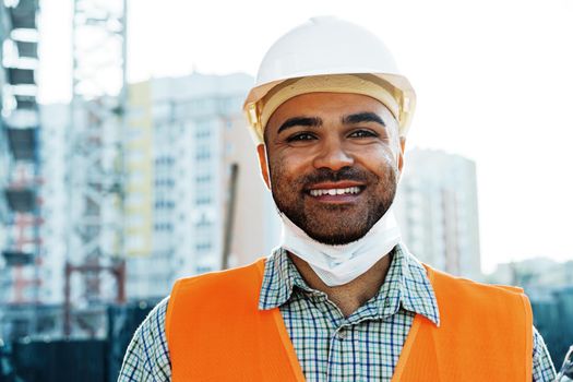 Portrait of mixed race man builder in workwear and hardhat wearing medical mask, close up photo