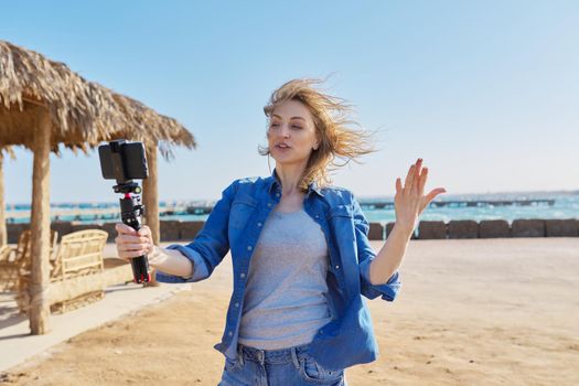 Middle aged woman looking at smartphone webcam talking recording video on sandy beach. Female blogger vlogger using video call on smartphone for blog, online stream, natural sea resort landscape