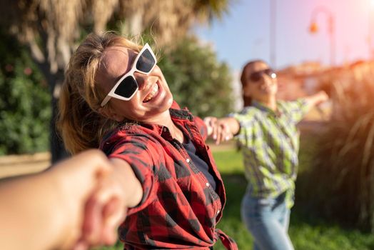 Pretty young caucasian woman in sunglasses pulling along by the hand invitingly with a happy smile as her friend on the background. Sunny summer day.