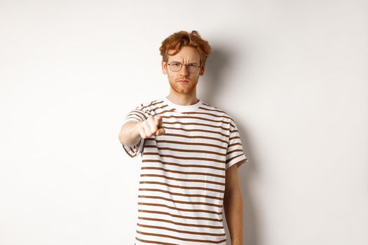 Angry and serious guy with red hair accusing you, frowning and pointing finger at camera, blame someone, standing over white background.