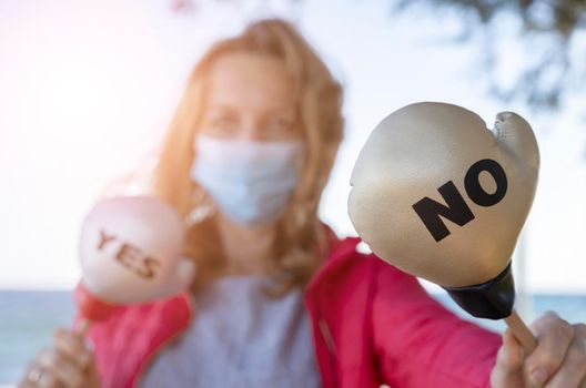 Young woman in protective sterile medical mask on sea background, holding small boxing glove with Yes No signs. America president election, pandemic coronavirus concept.