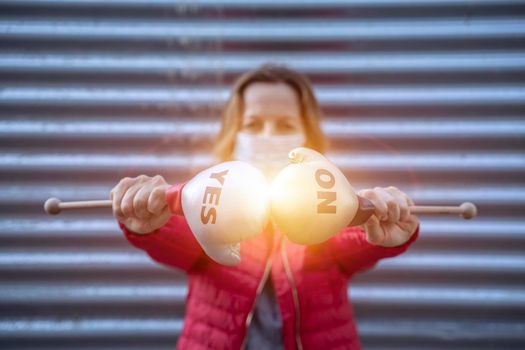 Young woman in protective sterile medical mask on her face, on striped background, holding small boxing gloves with Yes No signs. America president election, pandemic coronavirus concept.