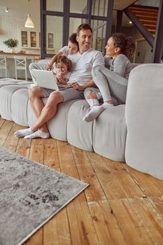 Low angle of cmiling parents sitting with daughter and son on sofa and watching laptop together