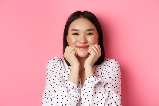 Beauty and lifestyle concept. Close-up of adorable asian woman showing puffy cheeks, smiling and looking happy, standing over pink background.