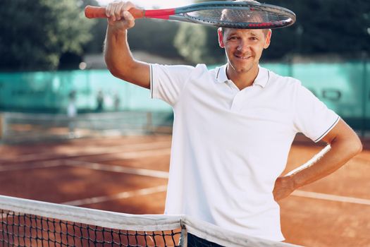 Portrait of positive male tennis player with racket standing at clay court near net