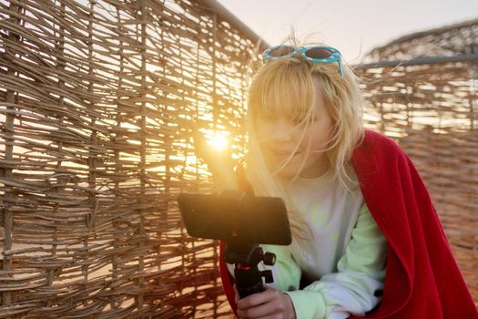 Outdoor sunset portrait of female teenager recording video on smartphone, rattan, sky background. Video call, stream, blog, vlog, telecommunications technologies, youth concept, copy space