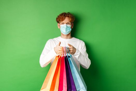 Pandemic and lifestyle concept. Cheerful redhead man going shopping in store, wearing medical mask and holding bags, standing over green background.