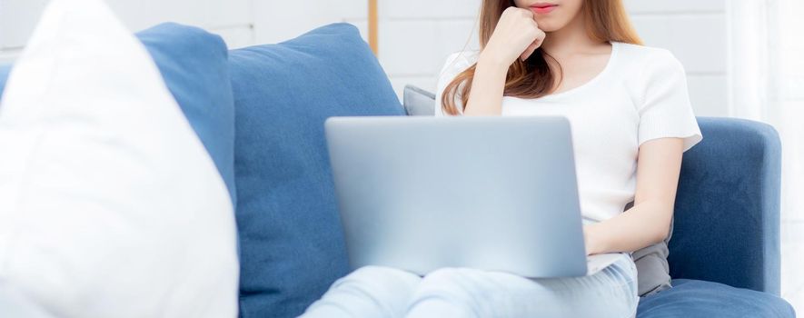 Young asian business woman smile and work from home with laptop computer online to internet on sofa in living room, freelance girl using notebook on couch with comfort, new normal, lifestyle concept.