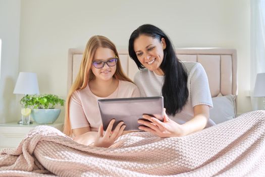 Happy mom and preteen daughter hugging together looking at screen of digital tablet, sitting at home in bed under blanket. Family, children, relationships, home, lifestyle, people concept