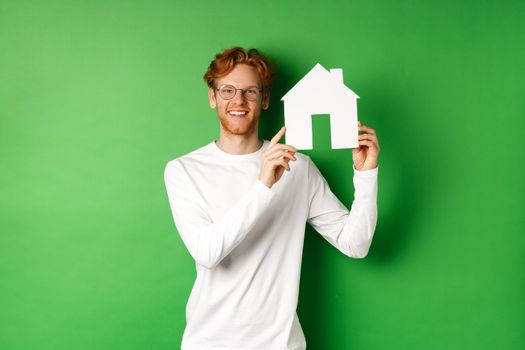 Real estate. Handsome young man with red hair, wearing glasses, showing paper house cutout and smiling, standing against green background.