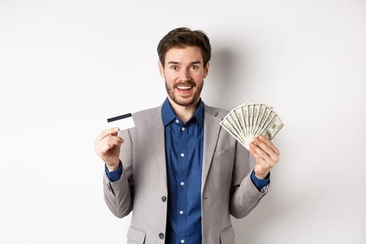 Happy successful businessman in suit showing money and credit card, smiling at camera, white background.