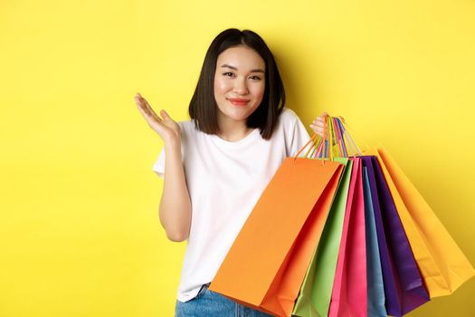 Silly asian girl showing shopping bags and shrugging, shop in stores with discounts, standing over yellow background.