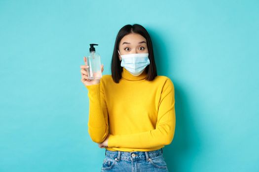 Covid-19, social distancing and pandemic concept. Cheerful asian woman showing hand sanitizer, recommend use antiseptic from coronavirus, wearing medical mask.