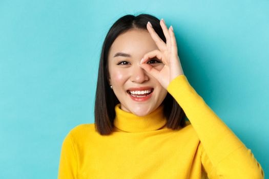 Beauty and makeup concept. Close up of carefree asian girl showing OK sign on eye and smiling, looking happy at camera, standing over blue background.