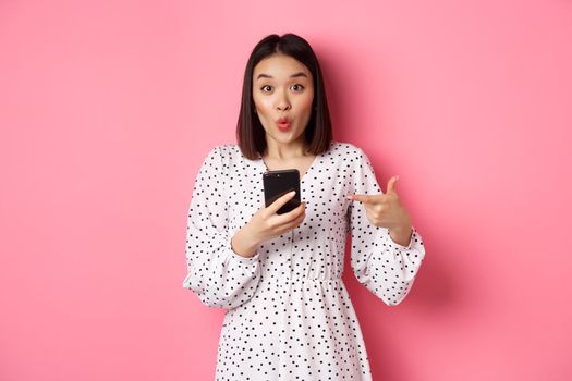 Online shopping and beauty concept. Amazed asian woman pointing finger at mobile phone, recommending smartphone app, standing over pink background.