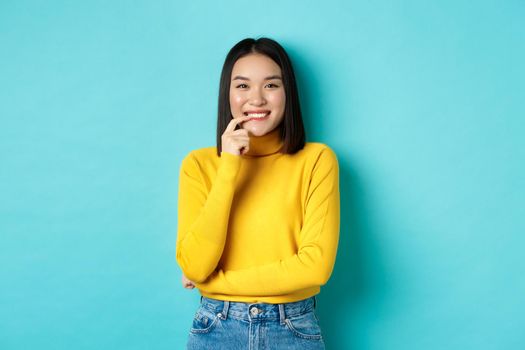 Image of good-looking asian woman in stylish outfit, touching lip and smiling at camera with happy face, standing over blue background.