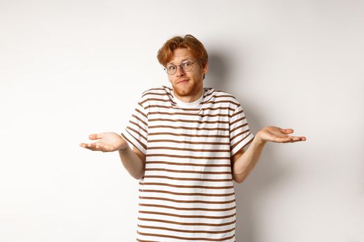 Confused young man with red hair, shrugging shoulders and looking indecisive, standing over white background.