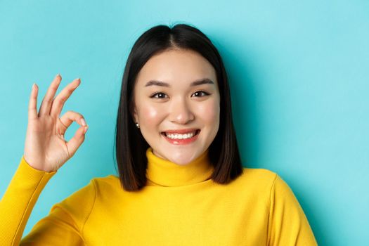 Beauty and makeup concept. Close up of happy asian woman with healthy shiny skin showing OK sign and smiling satisfied, standing over blue background.