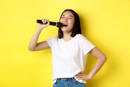 Happy asian woman singing song in karaoke, holding microphone, standing over yellow background.