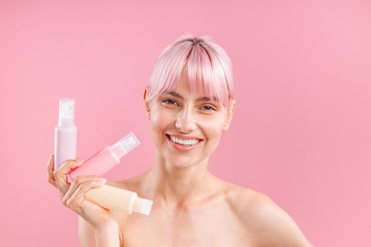 Happy young woman with pink hair holding three plastic travel bottles with beauty products, posing isolated over pink background. Beauty, spa, body care concept