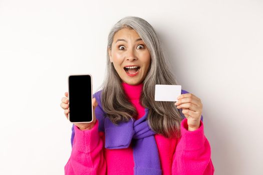 Online shopping. Closeup of amazed asian senior woman showing blank mobile screen and plastic credit card, looking happy, standing over white background.