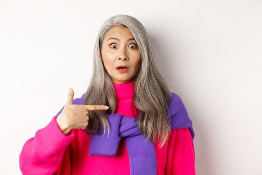 Confused asian senior woman with grey hair gasping startled, pointing at herself and looking at camera, standing over white background.
