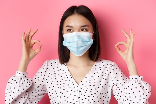 Covid-19, pandemic and lifestyle concept. Kawaii asian woman pointing fingers at her face mask, showing okay signs and looking left, standing over pink background.