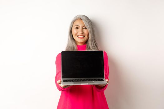 E-commerce concept. Smiling asian senior woman showing blank laptop screen and looking happy, demonstrating promo, white background.