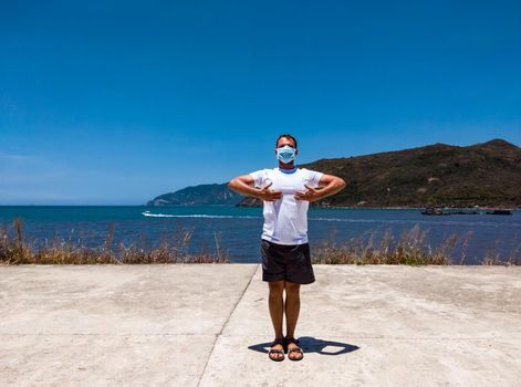 Coronavirus COVID 19. Young beautiful man doing sport performing workouts exercises in the park near sea during quarantine. Active life in surgical sterilizing face mask protection.