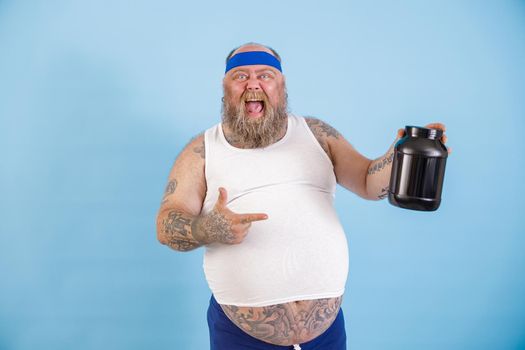 Joyful mature obese man with headband presents large bottle of nutrient supplement standing on light blue background in studio