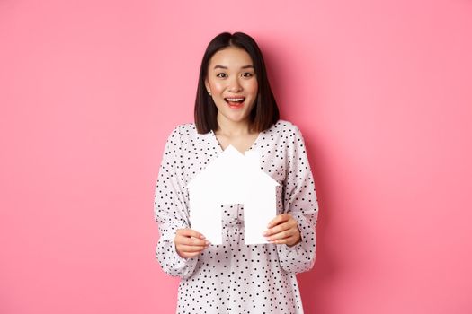 Real estate. Amazed asian woman searching for flat, holding house model and looking at camera happy, standing over pink background.