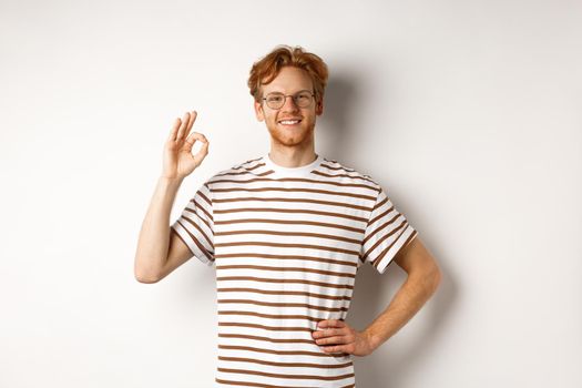 Confident smiling man with red hair assuring you, showing OK sign, guarantee quality, recommending something good, standing over white background.