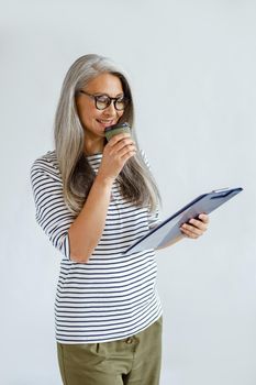 Smiling mature Asian lady with loose hair drinks coffee reading paper on clipboard standing on light grey background in studio