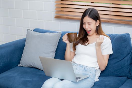 Young asian business woman smile and work from home with laptop computer online on sofa in living room, freelance girl using notebook and drink coffee on couch, new normal, lifestyle concept.