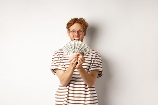 Shopping and finance concept. Lucky redhead guy winning, showing prize money and smiling happy, standing in glasses and t-shirt over white background.