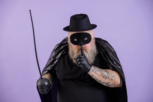 Middle aged bearded plump man in carnival Zorro costume with cape, mask and toy epee shows Hush gesture on purple background in studio