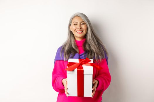 Beautiful asian senior woman smiling, congratulating with valentines day, holding gift in box, standing over white background.