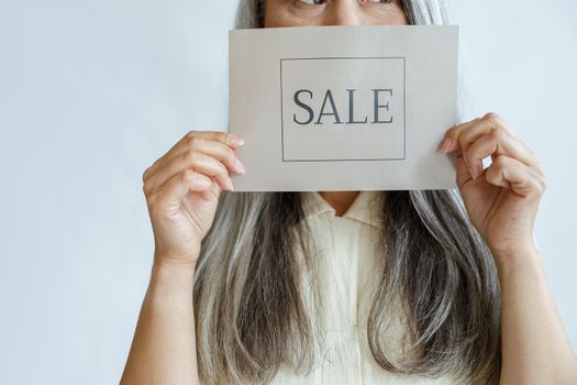 Middle aged lady with loose grey hair holds Sale sign near face standing on light background in studio closeup