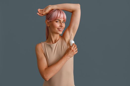 Happy young woman with pink hair in beige bodysuit smiling at camera while using antiperspirant, posing isolated over gray studio background. Beauty, body care concept