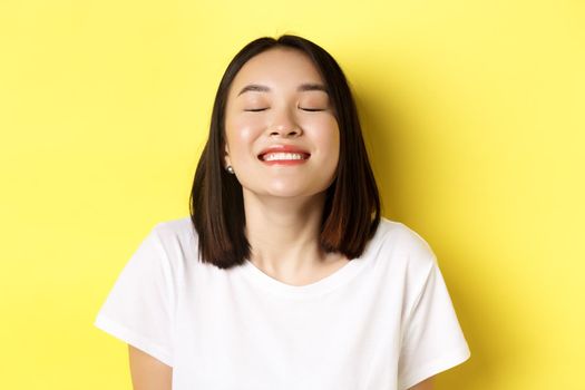 Close up of happy and relaxed asian woman enjoying sun, smiling with eyes closed and looking joyful, standing over yellow background.