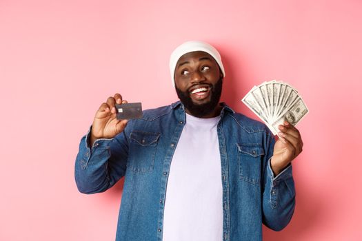 Dreamy african-american man showing credit card and dollars, thinking about shopping and smiling, standing over pink background.
