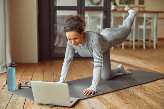 Cheerful slim woman is doing exercises on mat while watching training on laptop at home
