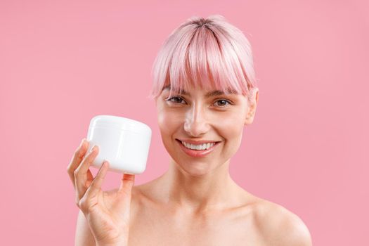 Portrait of smiling woman with pink hair holding white jar with nourishing body cream after shower isolated over pink background. Beauty, spa, body care concept