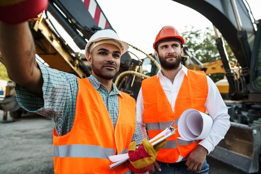 Two men engineers in workwear discussing their work standing against construction machines