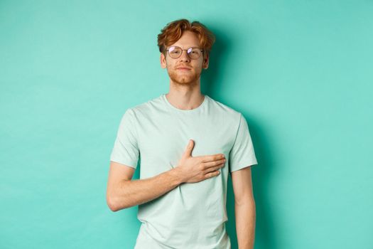 Proud young redhead man in glasses holding hand on heart, listening national anthem with respect, standing in t-shirt against turquoise background.