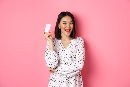 Shopping concept. Confident and happy asian woman holding credit card and smiling satisfied, standing against pink background.