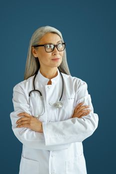 Skilled mature woman doctor in white uniform with glasses and crossed arms stands on blue background in studio. Professional medical staff